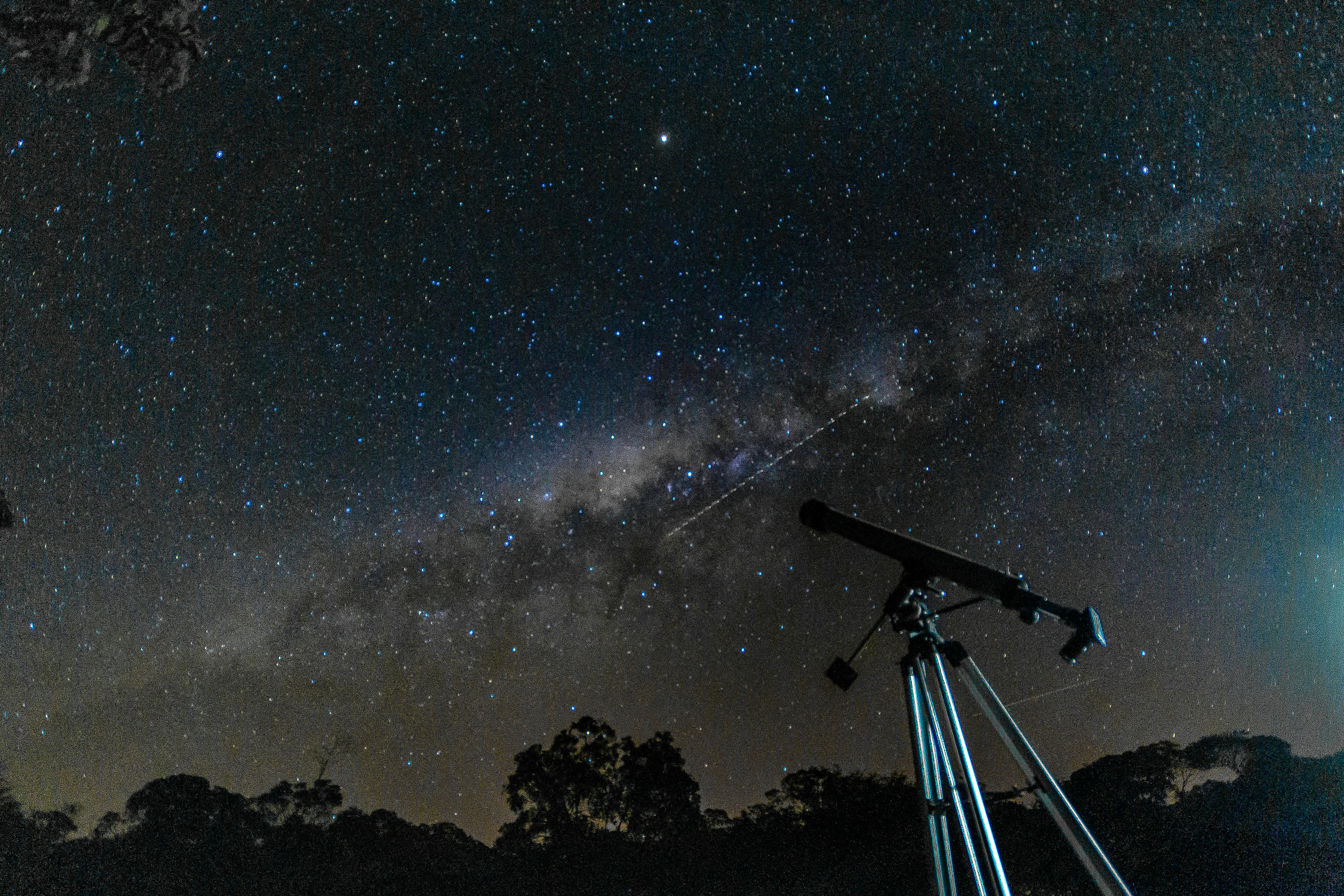 Tripod telescope silhouetted below the Milky Way above a dark treeline