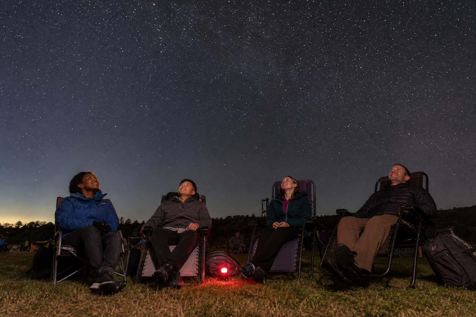 Four friends in reclining outdoor chairs look up at a star-filled sky during a community stargazing night.