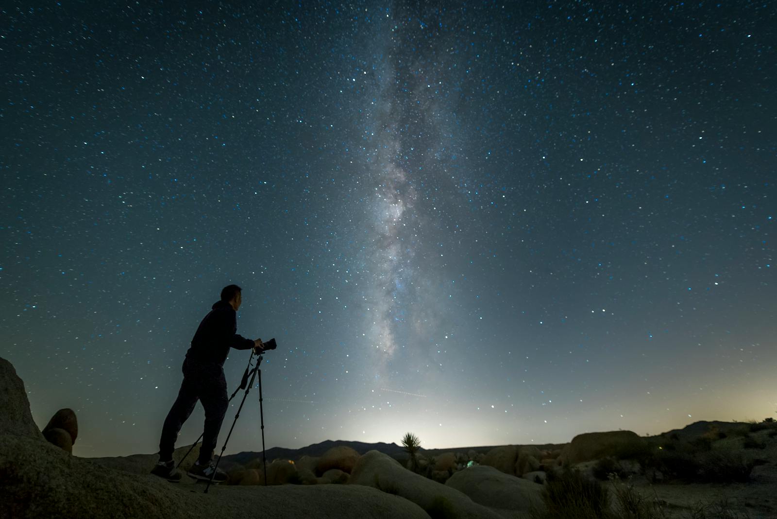 Photographer silhouetted against the Milky Way capturing night sky with DSLR on tripod