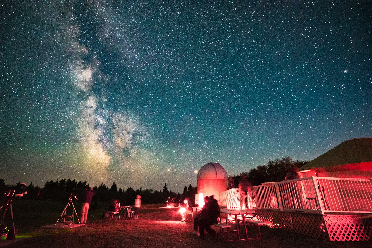 Astronomy community members gather beneath the Milky Way beside an observatory using red lights and telescopes